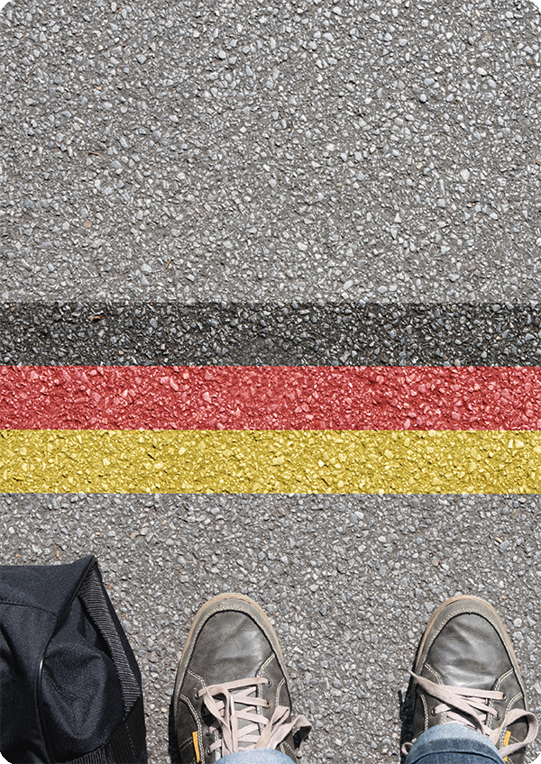 Foto aus der Perspektive einer Person, die auf ihre Schuhe herunterblickt und daneben eine Reisetasche; auf der Straße vor den Füßen der Person sind grafisch die Farben der Deutschlandfahne in Form einer Start oder Ziellinie gesetzt