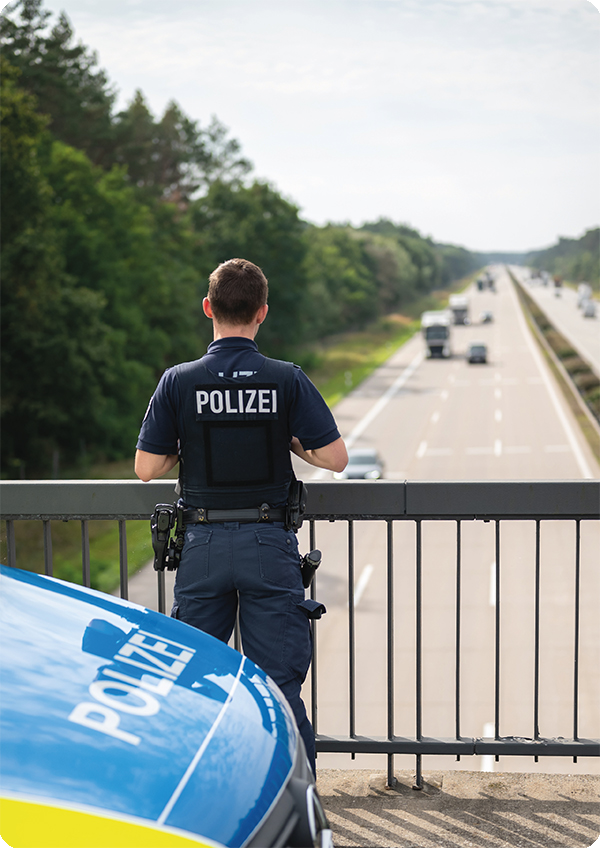 Foto eines Polizisten der mit dem Rücken zur Kamera auf einer Autobahnbrücke neben seinem Einsatzfahrzeug steht